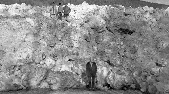 Quatre hommes posent pour la photo en noir et blanc devant une grosse avalanche qui recouvre une route.
