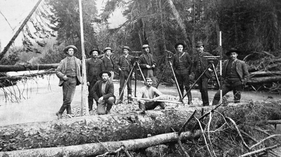 10 men with surveying equipment pose for black and white photo on snow covered ground in a forest.