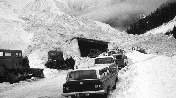 Photo en noir et blanc de plusieurs véhicules et plus de 3 chasse-neige stationnés à l'extérieur d'un pare-avalanche couvert de débris d'avalanche.