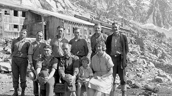 8 men, 1 woman and 1 child pose for black and white photograph with large sheds in background.