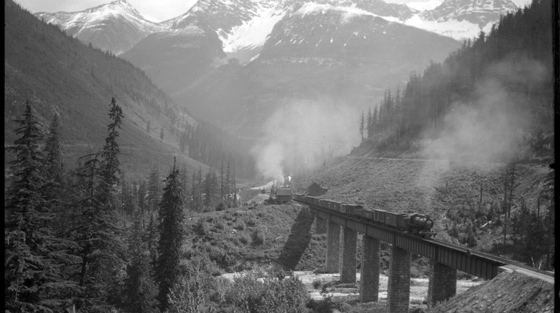 Black and white photograph of train crossing bridge in mountain pass.