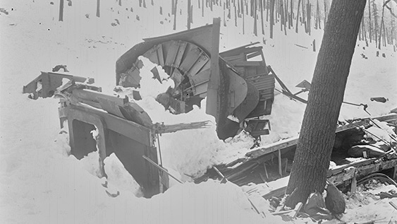 Black and white photograph of mangled rotary snow plough covered in avalanche debris.