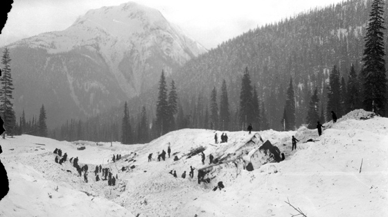 Black and white photograph of 20+ rescuers searching for victims in avalanche debris.