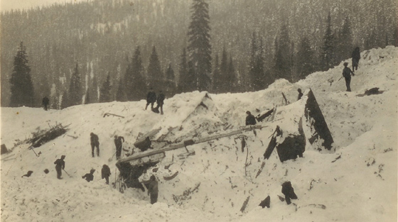 Close-up black and white photograph of 10+ rescuers digging through avalanche debris on slope.