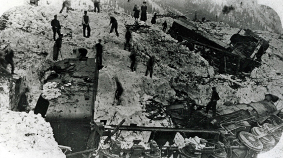 Une famille (au centre, en haut) observe plus de 10 hommes qui creusent pour déneiger un chasse-neige rotatif et une locomotive à vapeur ensevelis.