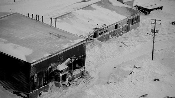 Black and white aerial photograph of two damaged buildings and avalanche debris.