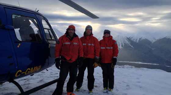 Trois secouristes vêtus de manteaux rouges posent pour la photo en couleur sur le sommet d'une montagne, devant un hélicoptère bleu de recherche et de sauvetage.