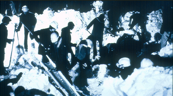 Black and white close-up photograph of up to 6 rescuers with shovels on site of avalanche.