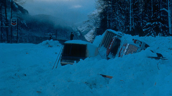 Photographie en couleur d'un teint bleuté montrant deux véhicules renversés, couverts de débris d'avalanche dans une vallée montagneuse.