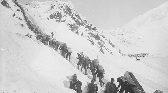 Black and white photograph of dozens of men with packs hiking in line up steep, snow-covered mountain slope.