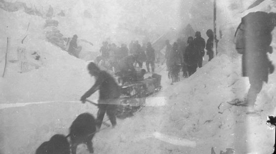 Photographie en noir et blanc d'une vue rapprochée de près d'une centaine d'hommes et de plusieurs chiens tirant des traîneaux en bas d'une montagne escarpée en pleine tempête de neige. 