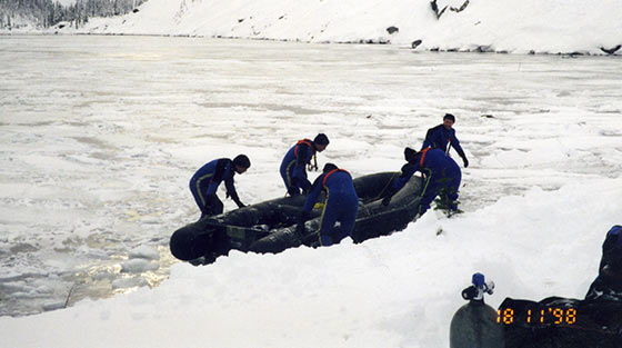 Photographie en couleur de 5 membres d'une équipe de plongée policière qui manoeuvre un bateau pneumatique noir sur un lac gelé.