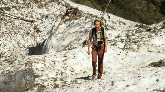 Photo en couleur d'un homme qui se promène sur la zone d'arrêt enneigée d'un couloir d'avalanche dans une vallée en été.