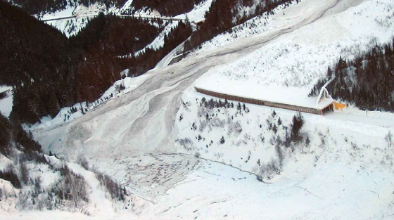 Aerial, colour photograph of avalanche partially blocking river and highway in mountain pass.